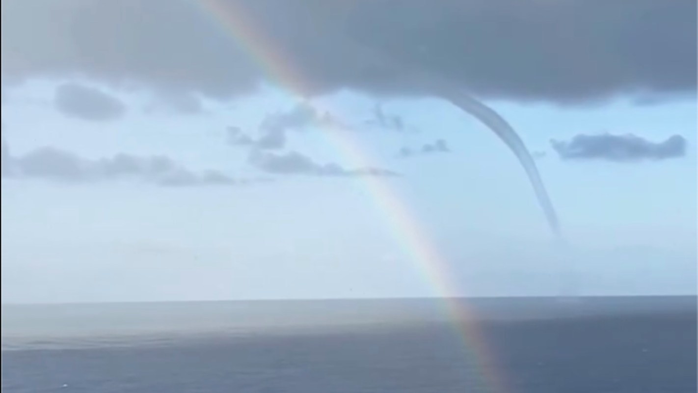 A waterspout forms over the ocean under a cloudy sky, with a rainbow arcing across the scene.