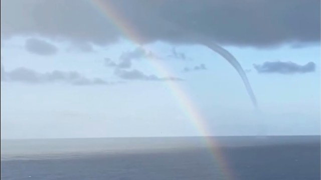 A waterspout forms over the ocean under a cloudy sky, with a rainbow arcing across the scene.
