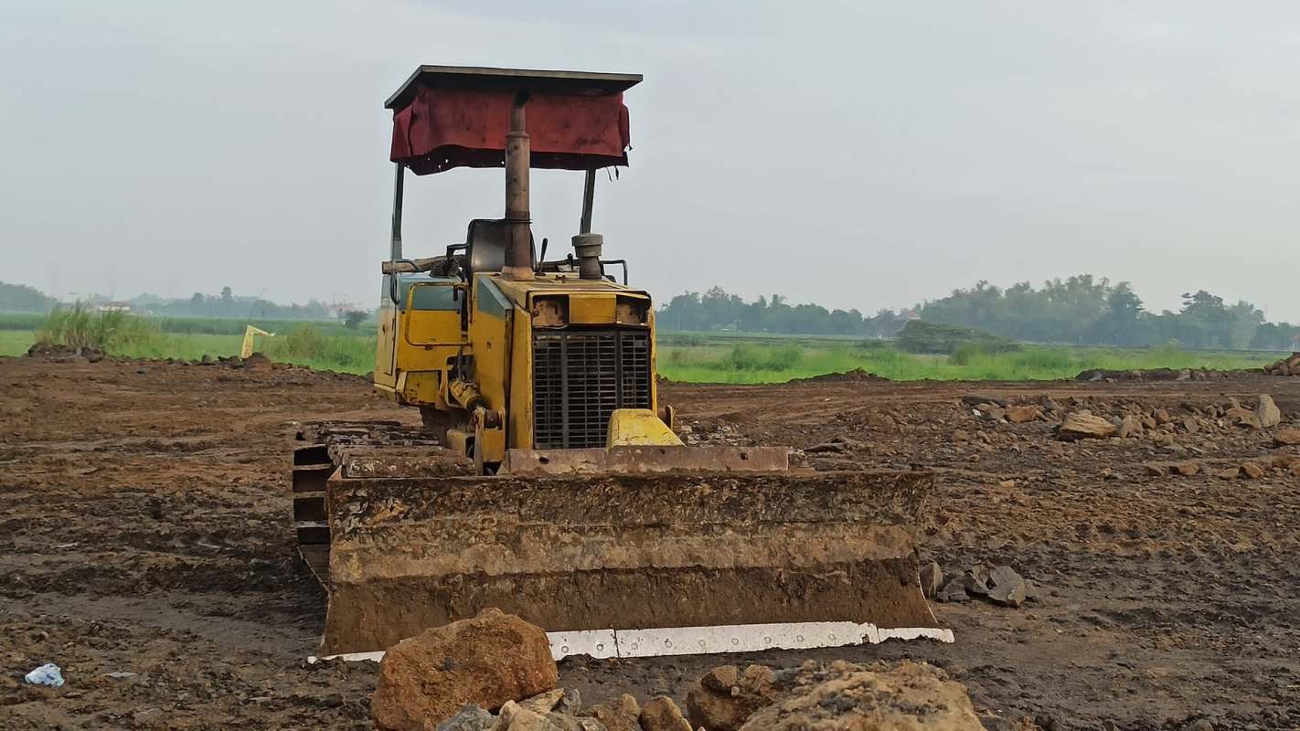 A yellow bulldozer with a red canopy is parked on a dirt site amid greenery and scattered rocks.