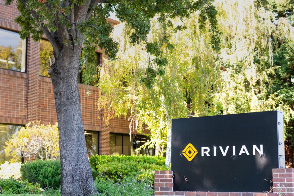 A black sign with the logo and name "RIVIAN" in front of a brick building surrounded by greenery.