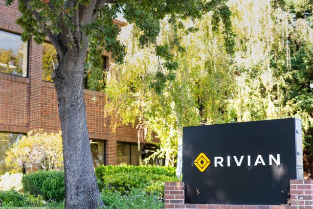 A black sign with the logo and name "RIVIAN" in front of a brick building surrounded by greenery.