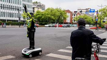 A robotic traffic officer signals at a busy intersection while a man on a bicycle watches from the sidewalk.