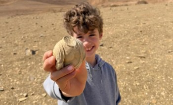 A smiling boy holds a round stone while standing in a dry, rocky landscape.