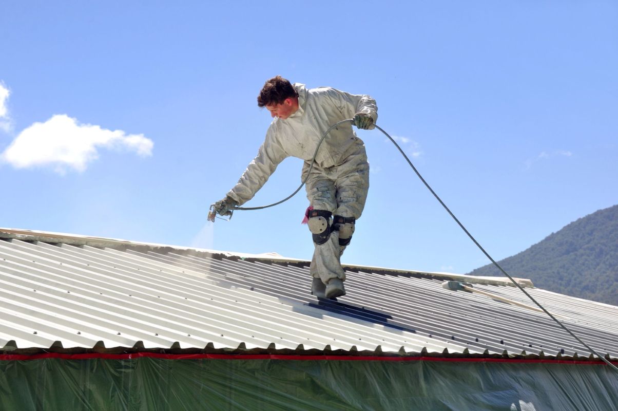 A person in protective gear spray paints a metal roof under a clear blue sky.