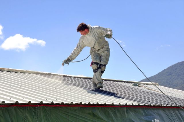 A person in protective gear spray paints a metal roof under a clear blue sky.