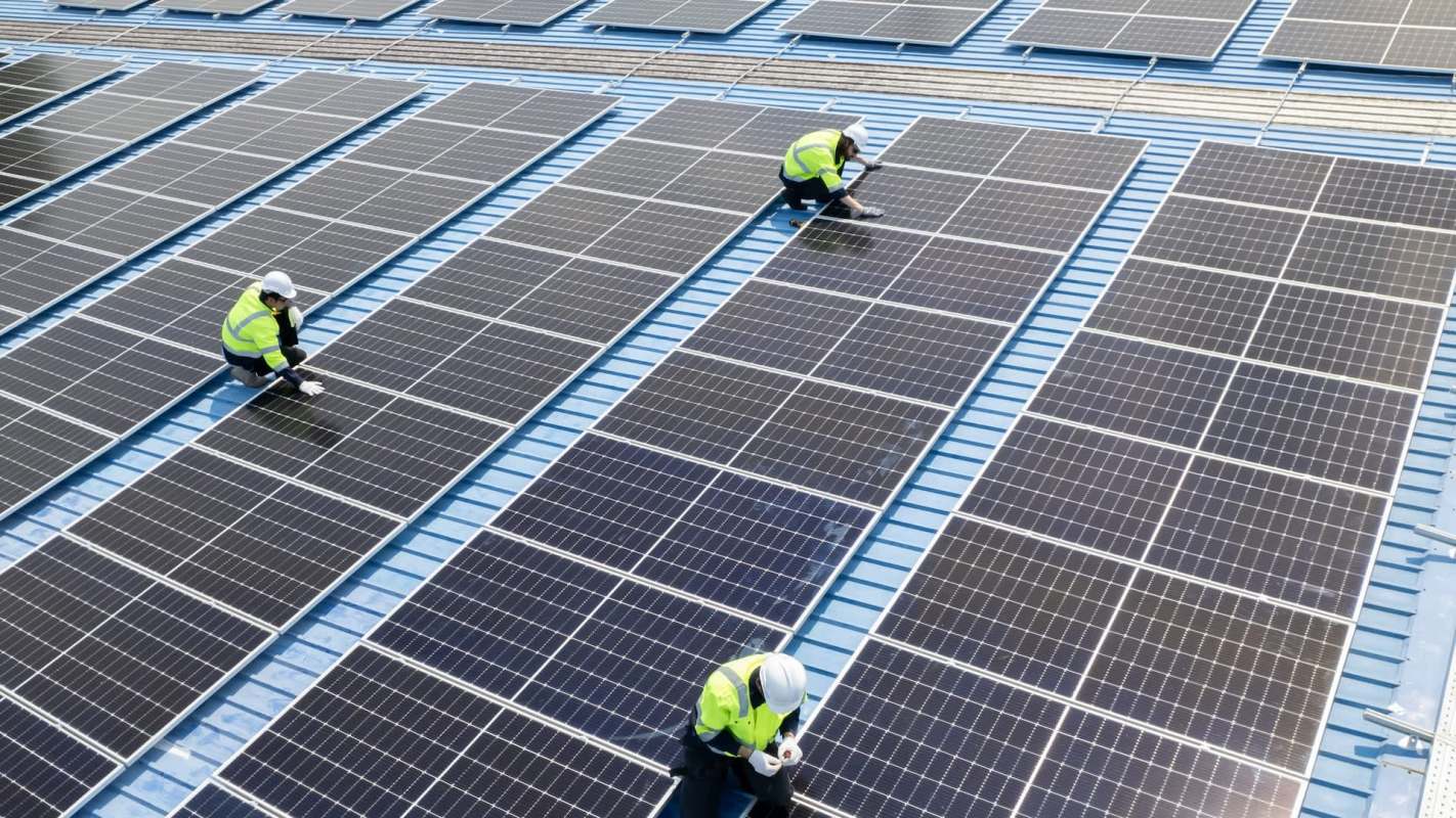 Technicians in safety vests work on a solar panel installation on a blue rooftop.