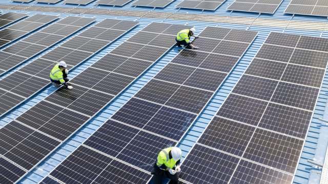 Technicians in safety vests work on a solar panel installation on a blue rooftop.