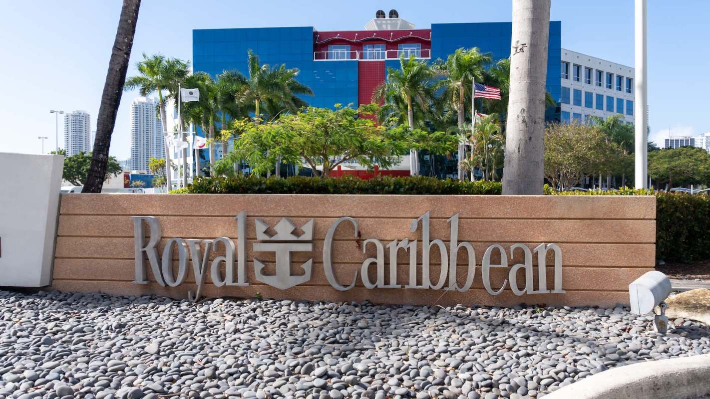 A sign for Royal Caribbean outside a modern building with palm trees and a clear blue sky in the background.