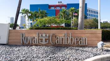 A sign for Royal Caribbean outside a modern building with palm trees and a clear blue sky in the background.