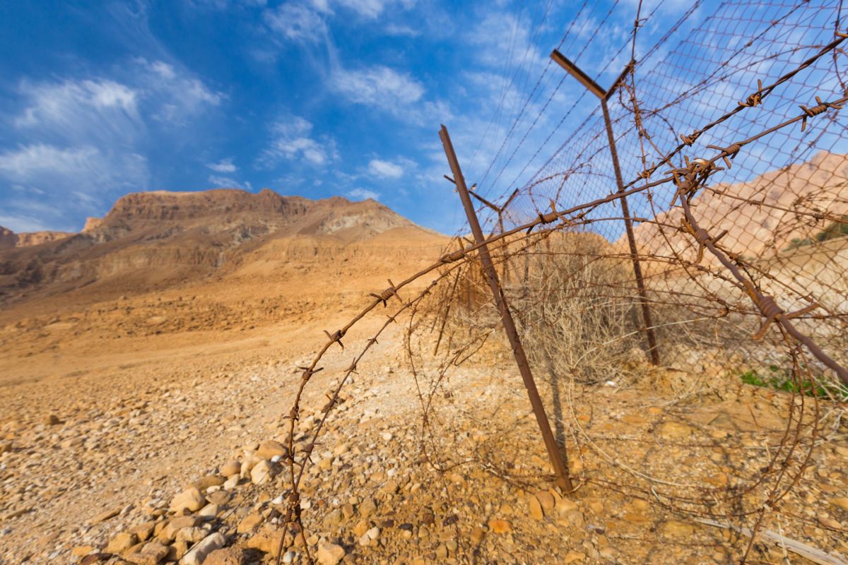 A barbed wire fence stretches across a rocky, arid landscape with mountains under a blue sky.