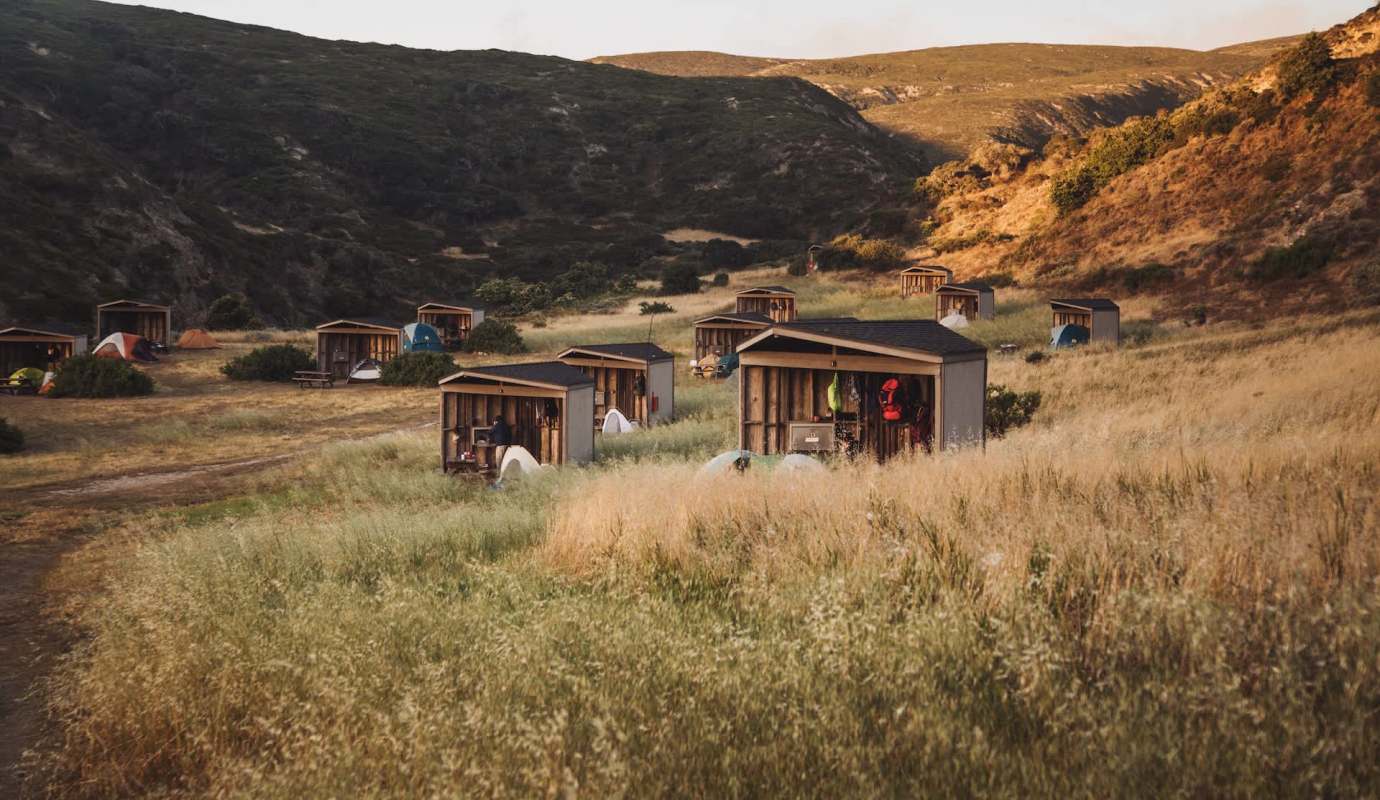 A serene campsite with several wooden cabins and tents amid tall grass and rolling hills.