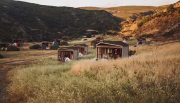 A serene campsite with several wooden cabins and tents amid tall grass and rolling hills.