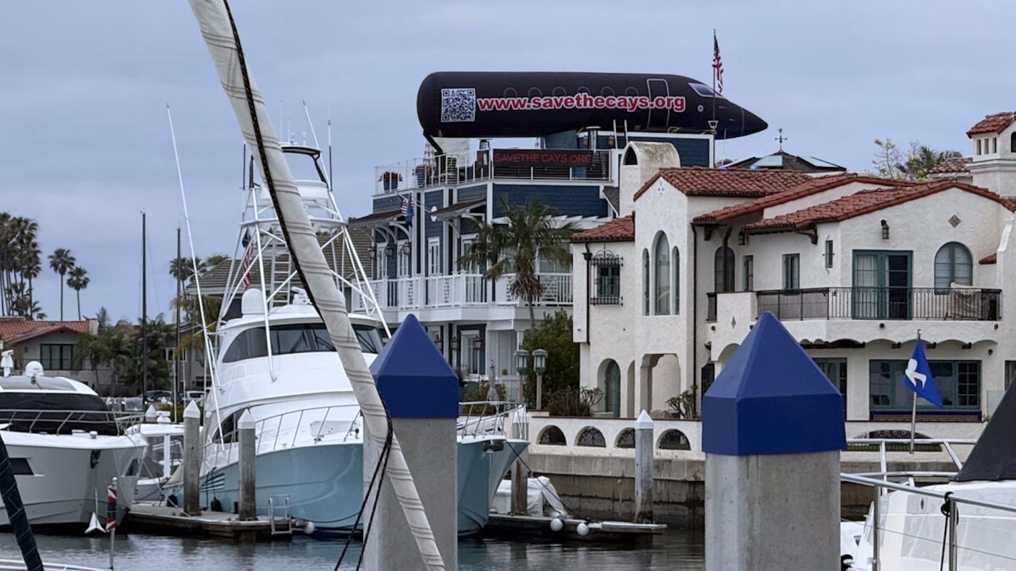 A floating billboard advertising "Save the Cays" on a house overlooking a marina with yachts and palm trees.