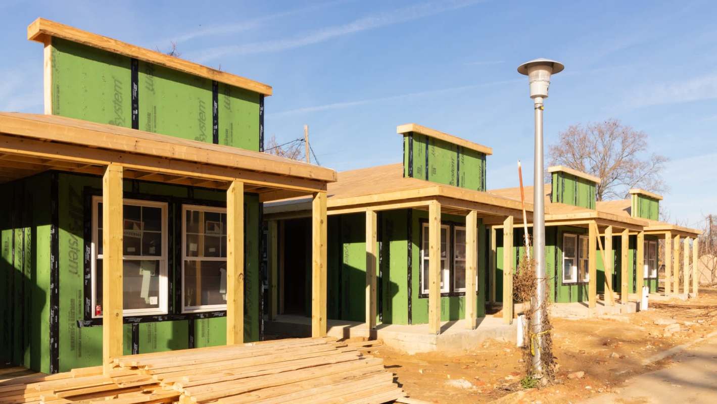 A row of new construction tiny houses with green siding and wooden supports under clear blue skies.
