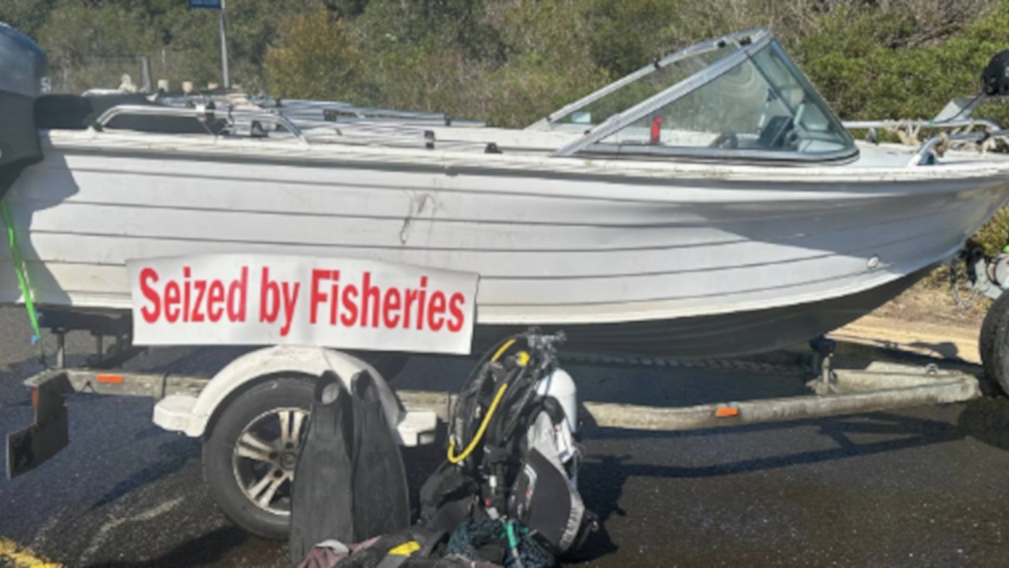 A small white boat on a trailer with a sign reading "Seized by Fisheries" and diving gear nearby.