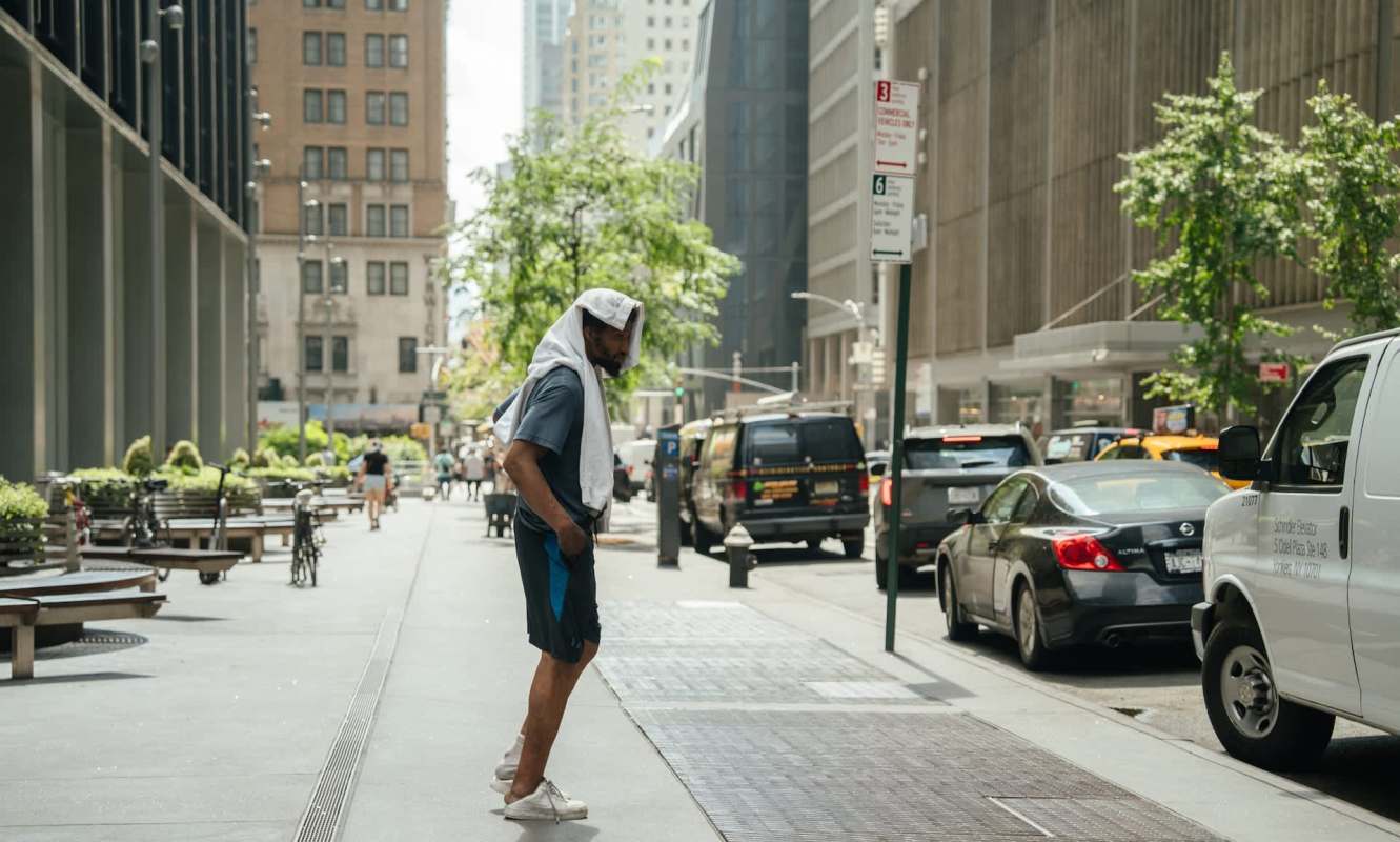 A man wearing a towel on his head stands on a city sidewalk, surrounded by parked cars and greenery.