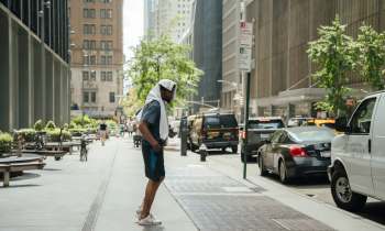 A man wearing a towel on his head stands on a city sidewalk, surrounded by parked cars and greenery.