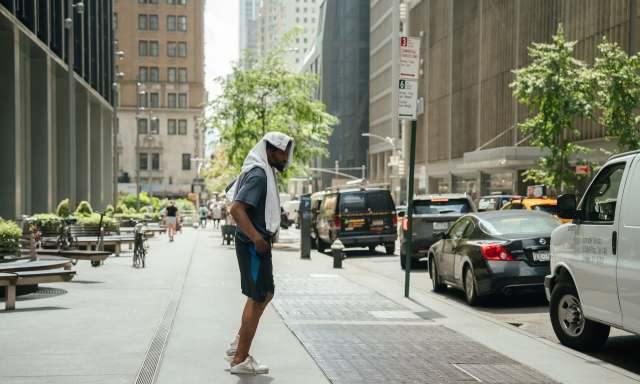 A man wearing a towel on his head stands on a city sidewalk, surrounded by parked cars and greenery.