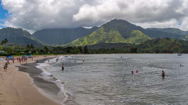 People walk and swim at a beach surrounded by tropical foliage and mountains.