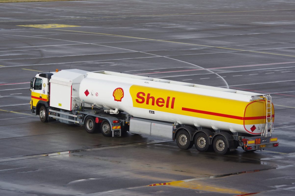 A Shell-branded fuel truck is parked on an airport tarmac, reflecting puddles on the wet surface.