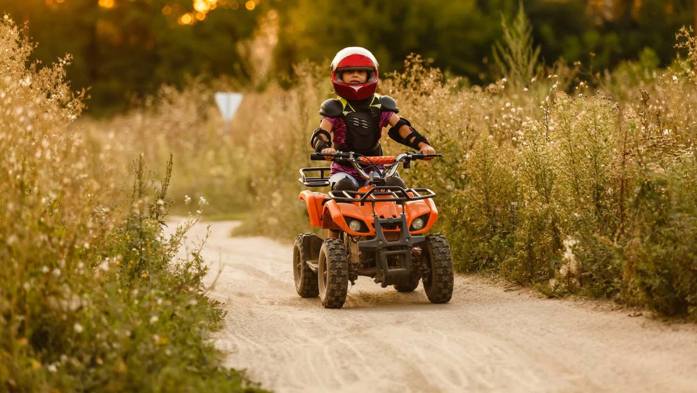A child in protective gear rides an orange ATV along a dirt path surrounded by tall grass and wildflowers.