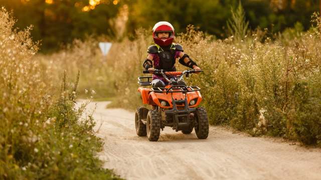 A child in protective gear rides an orange ATV along a dirt path surrounded by tall grass and wildflowers.