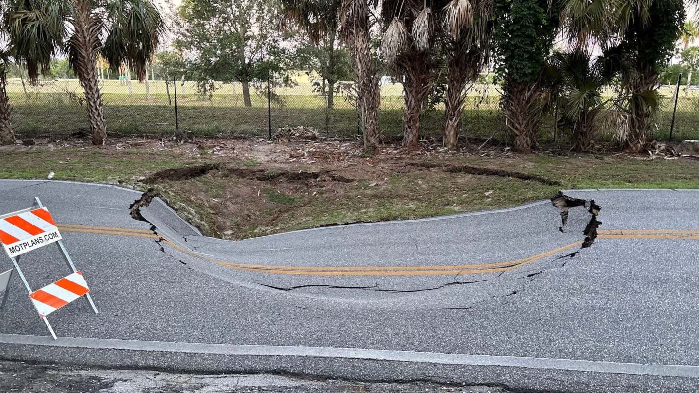 A cracked road with a noticeable dip, surrounded by palm trees and caution signs.