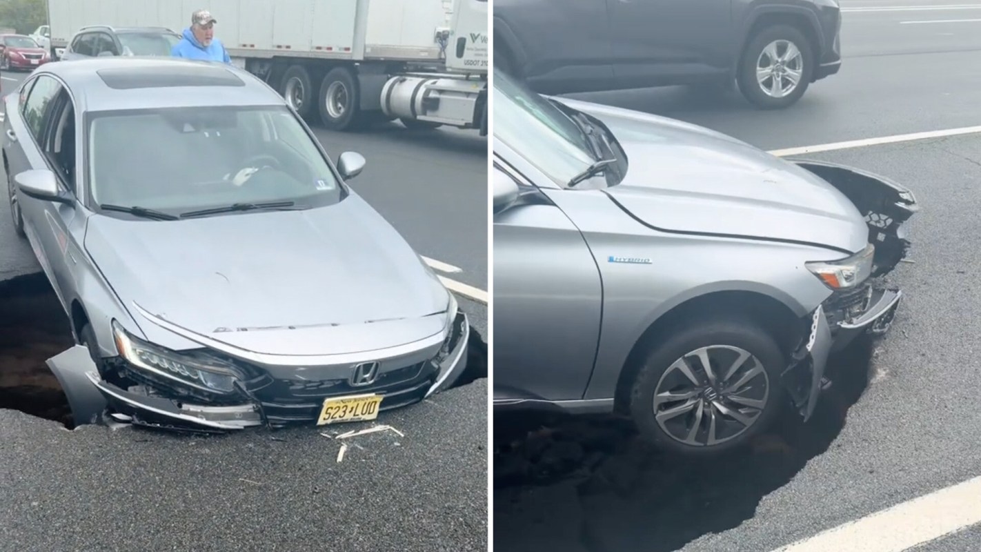 A silver car partially trapped in a large pothole on a highway, with visible front-end damage.