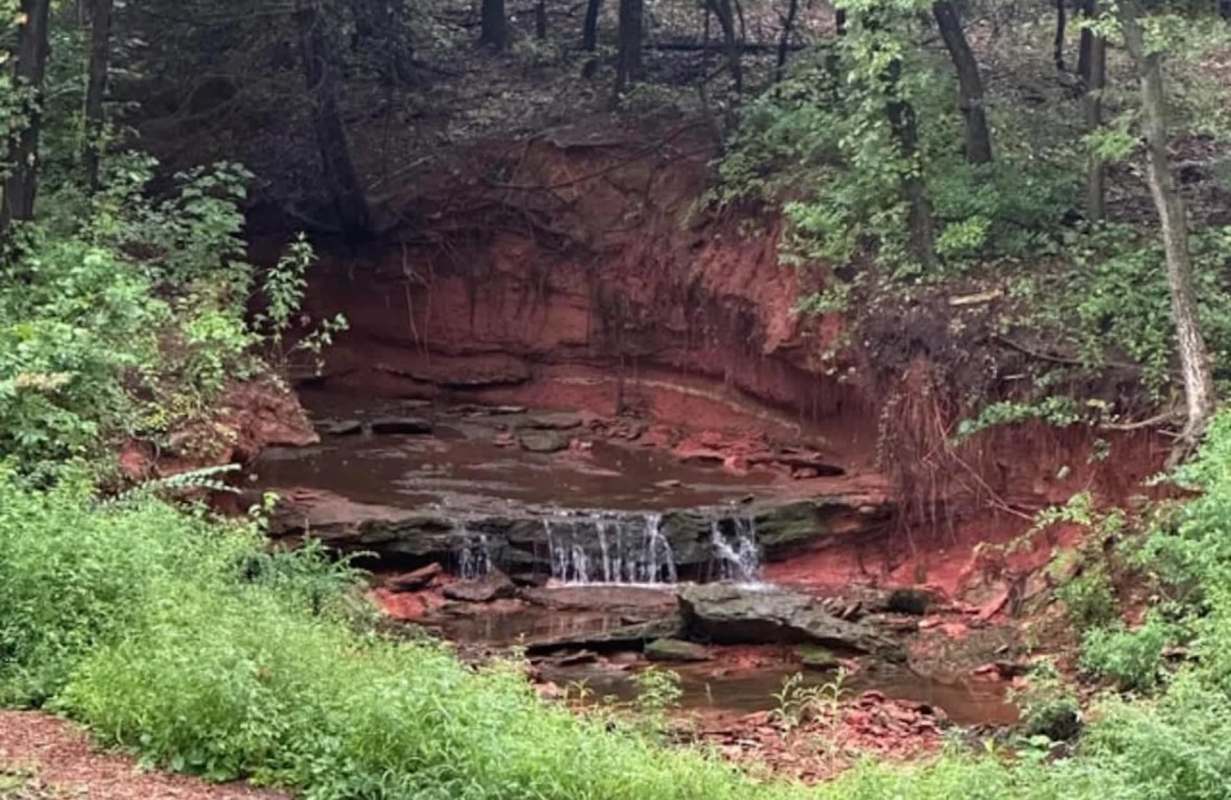 A small waterfall cascading over rocks surrounded by lush greenery and red clay banks in a wooded area.