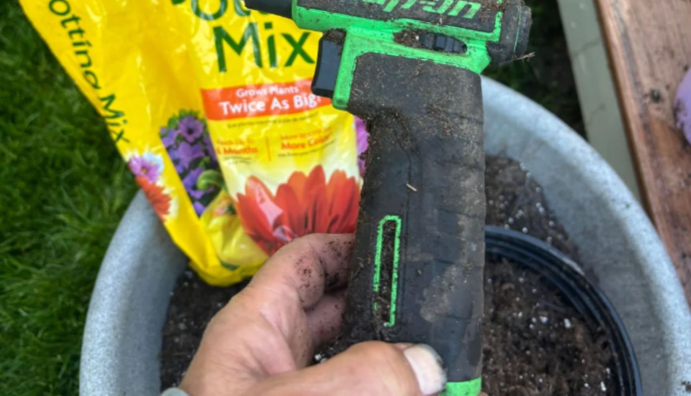A close-up of a hand holding a dirty gardening tool with a yellow potting mix bag in the background.
