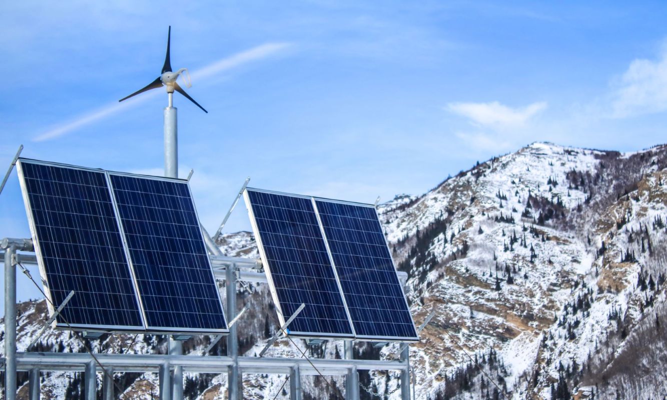A wind turbine and solar panels are positioned in front of a snow-covered mountain landscape under a blue sky.