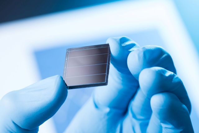 A gloved hand holds a small solar cell against a soft blue background.