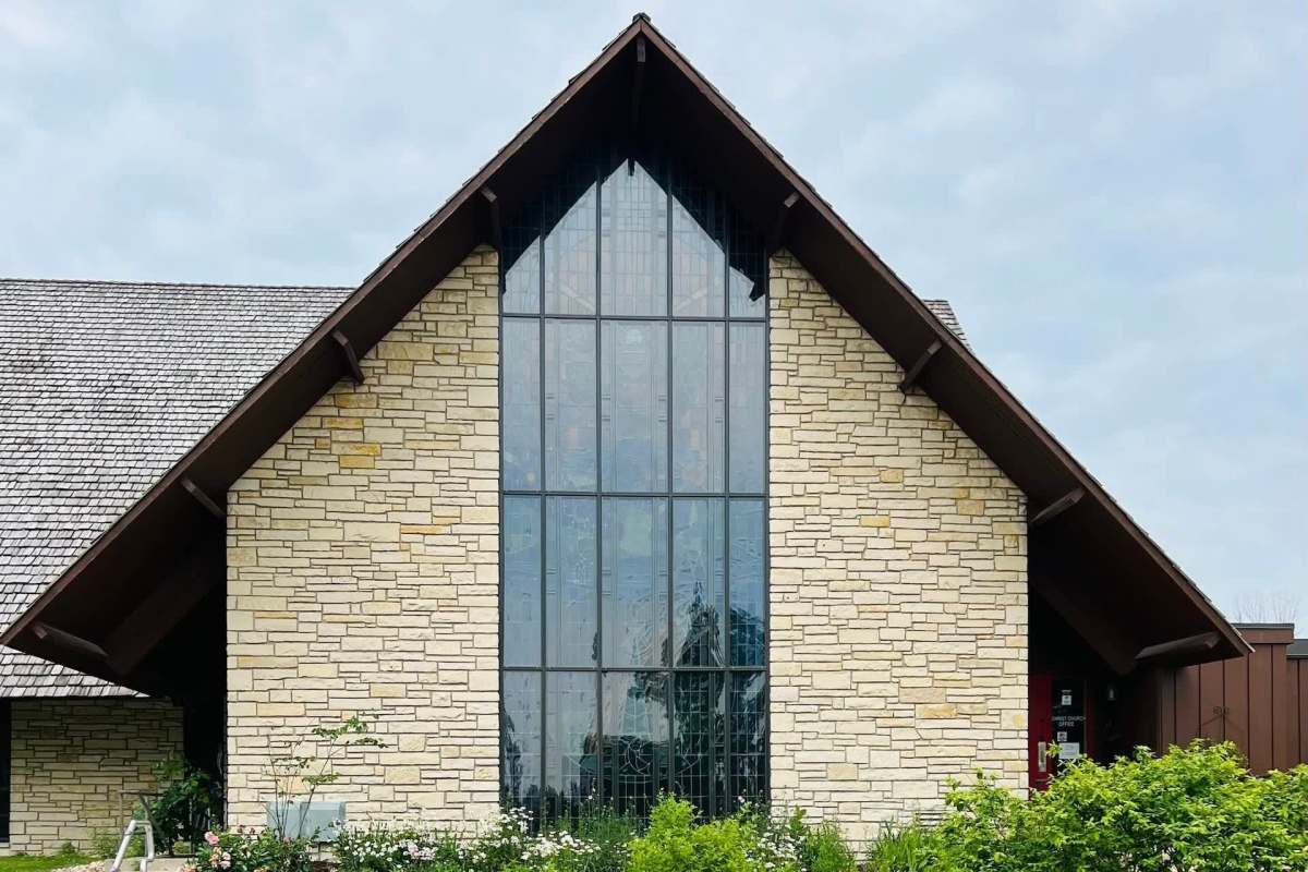 A modern stone church with a tall, glass-paneled front and a sharply sloped roof surrounded by greenery.