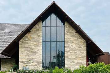 A modern stone church with a tall, glass-paneled front and a sharply sloped roof surrounded by greenery.