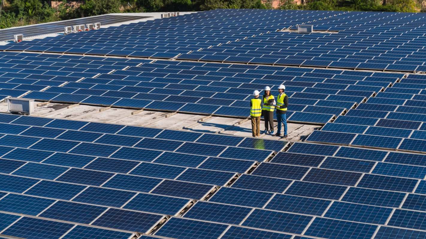 Three workers in safety vests discuss a project on a rooftop covered with solar panels.
