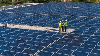 Three workers in safety vests discuss a project on a rooftop covered with solar panels.