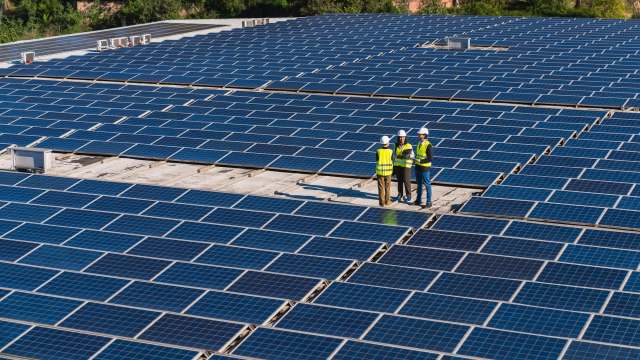 Three workers in safety vests discuss a project on a rooftop covered with solar panels.