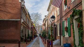 A narrow, brick-lined alley adorned with festive wreaths and flags on historic buildings under a cloudy sky.
