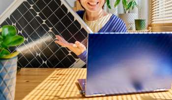 A person smiles while gesturing with a solar panel, sitting at a table with a laptop and potted plants in a sunlit room.