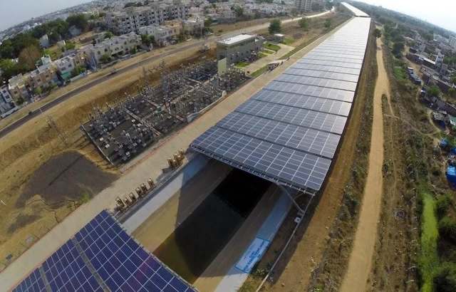 Aerial view of solar panels installed over a water canal, with electrical infrastructure nearby and residential buildings in the background.