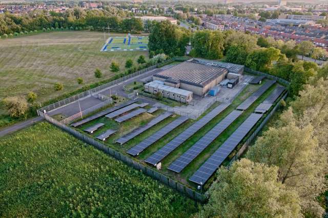 An aerial view of a building surrounded by solar panels and a grassy field with trees in the background.