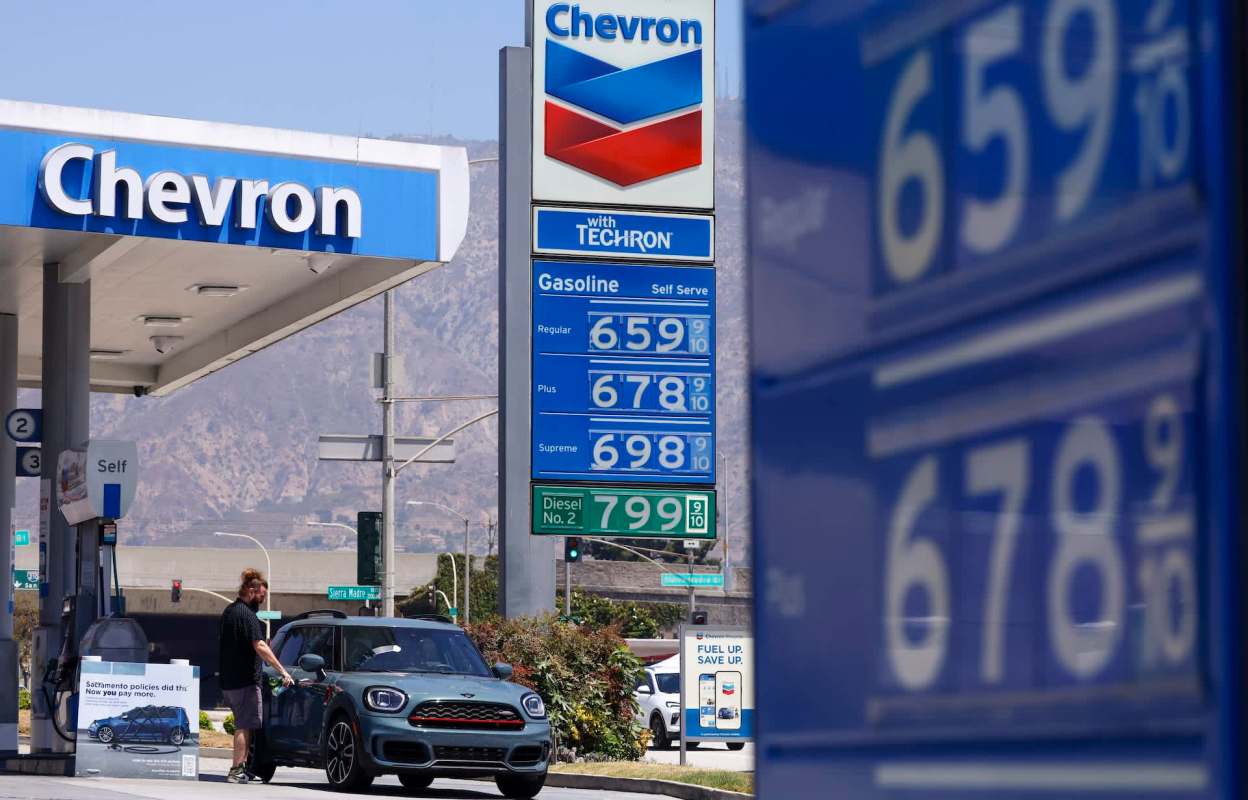 A person refuels a car at a Chevron gas station with fuel prices displayed in the background.