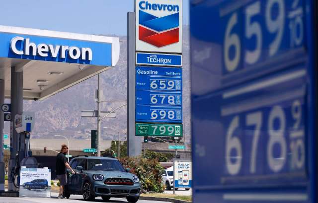 A person refuels a car at a Chevron gas station with fuel prices displayed in the background.