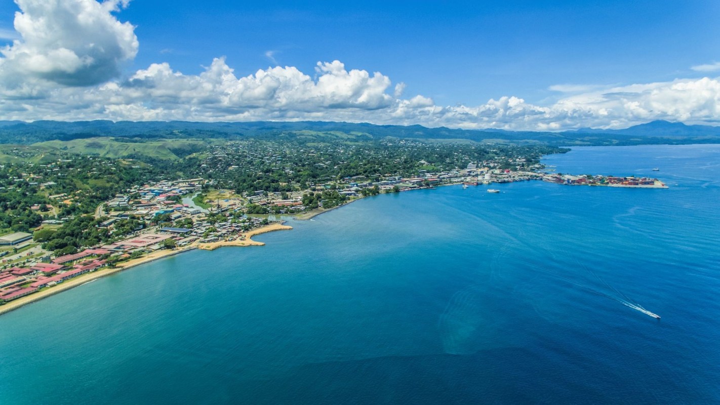 Aerial view of a coastal town with blue waters, green hills, and scattered clouds under a bright sky.