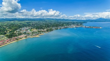 Aerial view of a coastal town with blue waters, green hills, and scattered clouds under a bright sky.