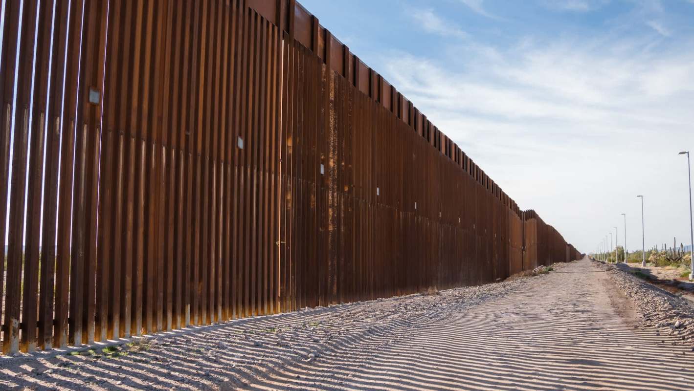 A rust-colored border wall stretches along a sandy path.