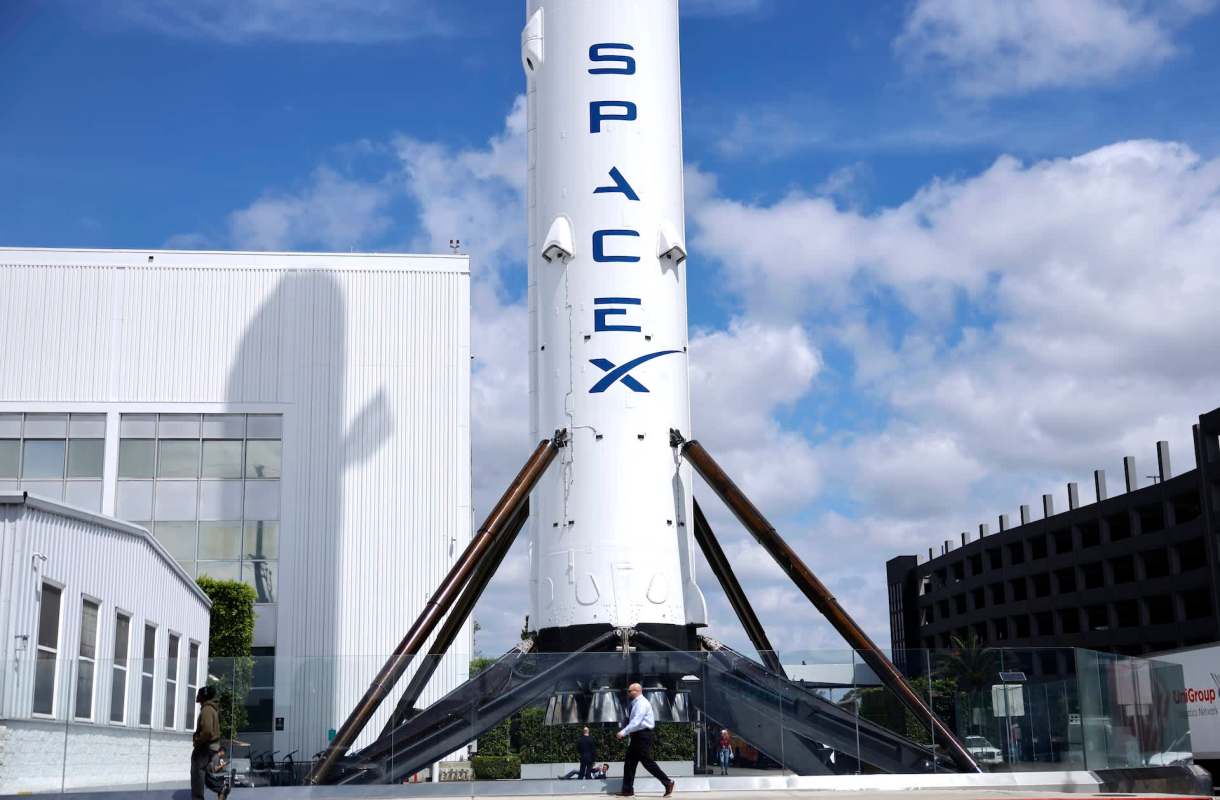 A SpaceX rocket stands tall on a sunny day as people walk below.