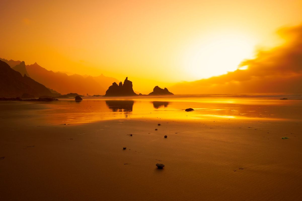 A golden sunset reflects on a calm beach with silhouetted rock formations in the distance.