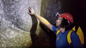 A climber in a helmet touches a textured rock surface in a dimly lit cave.
