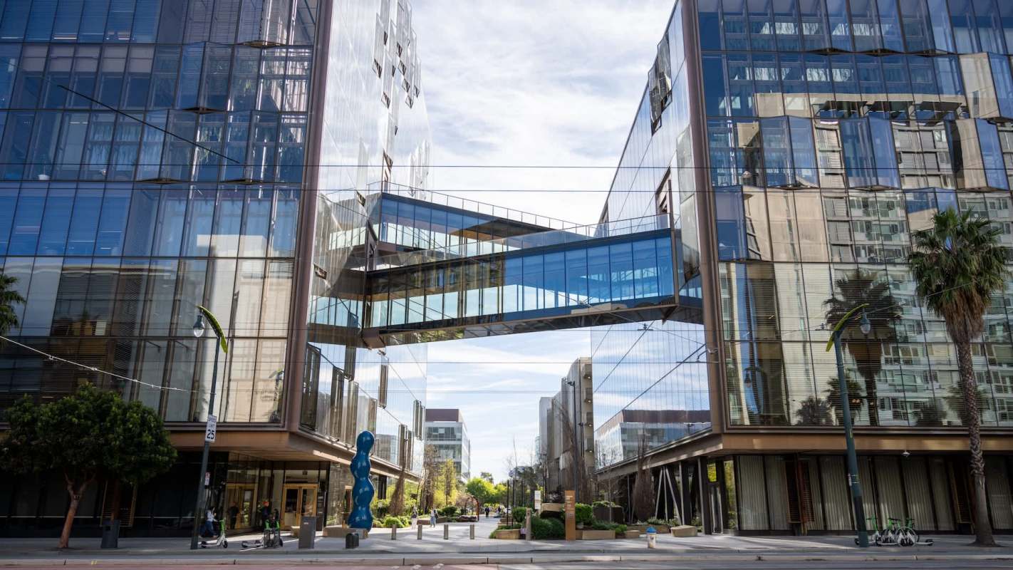 A modern urban plaza featuring glass buildings, a suspended bridge, and palm trees under a blue sky.